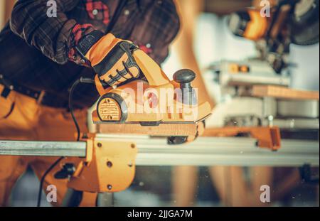 Holzbearbeitung auf der Baustelle. Professioneller Zimmermann, der die Holzplanke mit elektrischer Hobel-Maschine verarbeitet. Thema Industrieausrüstung. Stockfoto