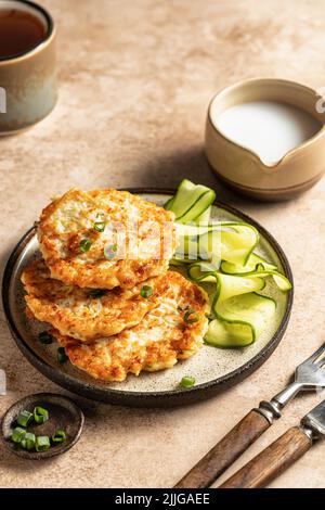 Zucchini-Pfannkuchen mit grüner Zwiebel und Gurke serviert mit Sahnesauce auf braunem Hintergrund mit Besteck und Tee. Vegetarisches Speisekonzept. Vertikal oder Stockfoto