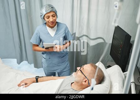 Horizontale Aufnahme aus einem hohen Winkel, bei der junge Erwachsene Frauen im Krankenhaus mit einem digitalen Tablet arbeiten, das dem reifen Mann im Bett etwas erzählt Stockfoto