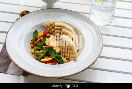 Hummus mit Tomaten und Kräutern auf einem weißen Teller mit Pita auf dem Tisch und einem Glas Wasser im Café vegan Stockfoto