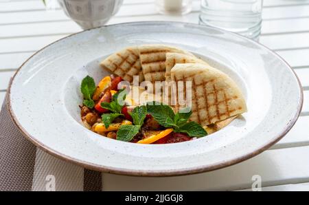 Hummus mit Tomaten und Kräutern auf einem weißen Teller mit Pita auf dem Tisch und einem Glas Wasser im Café vegan Stockfoto