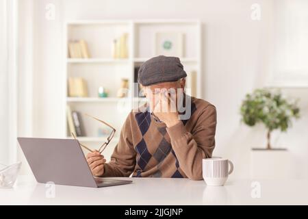 Älterer Mann, der Kopfschmerzen hat, ein Dokument hält und zu Hause vor einem Laptop sitzt Stockfoto