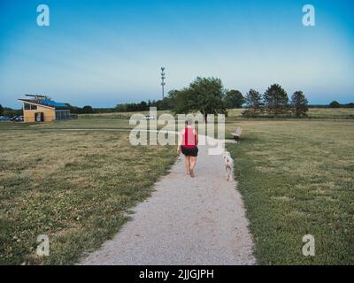 Eine Frau in Rot, die während der blauen Stunde einen cremefarbenen Standardpudel läuft. Foto aufgenommen im Heritage Park in Olathe Kansas. Stockfoto