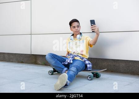 Kaukasischer Teenager, der auf einem Skateboard sitzt und mit einem Smartphone ein Foto von sich macht. Urbane Lebensart. Platz für Text. Stockfoto