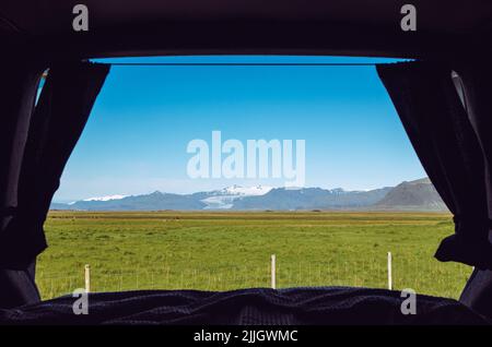 Blick aus dem Camper Minivan zurück aus dem eingebauten Bett. Reisen mit dem Van nach Island. Blick über Berge, Weide und Gletscher, blauer Himmel, sonniger Tag. Stockfoto