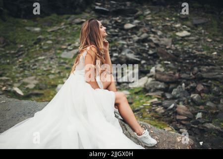 Braut in einem weißen Kleid posiert, sitzt auf einem Stein vor dem Hintergrund der felsigen Berge. Hochzeitsfoto von einem schönen kaukasischen Mädchen. Hochzeitsreise. Stockfoto