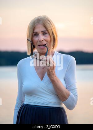 Fröhliche Senior fit elegante Dame in weiß und blau gekleidet mit Sonnenbrille genießen die Natur im Sommer bei Sonnenuntergang Stockfoto