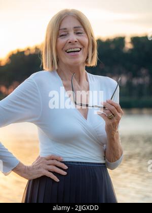 Fröhliche Senior fit elegante Dame in weiß und blau gekleidet mit Sonnenbrille genießen die Natur im Sommer bei Sonnenuntergang Stockfoto