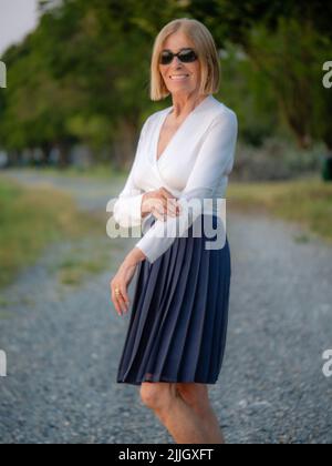 Fröhliche Senior fit elegante Dame in weiß und blau gekleidet mit Sonnenbrille genießen die Natur im Sommer bei Sonnenuntergang Stockfoto