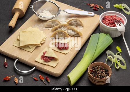 Rohe Gyoza-Knödel und gefüllte Teigstücke auf Schneidebrett. Fleischfüllung in Schüssel, Piment und Lauch. Draufsicht. Schwarzer Hintergrund Stockfoto