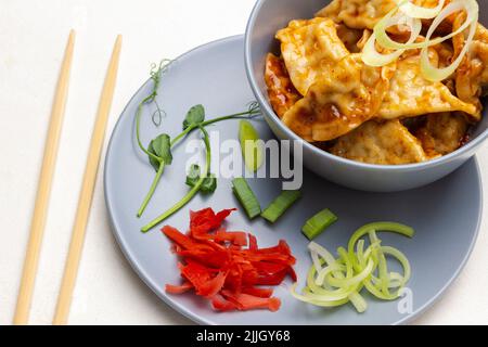 Essstäbchen auf dem Tisch. Gyoza-Knödel in grauer Schüssel. Grüne Erbsensprossen, Lauch und eingelegter Ingwer auf grauem Teller. Flach liegend. Weißer Hintergrund Stockfoto