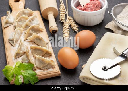 Rohe Gyoza-Knödel auf Schneidebrett. Teig- und Teigschneider, Eier und Nudelholz auf dem Tisch. Draufsicht. Schwarzer Hintergrund Stockfoto