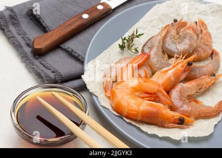 Garnelen auf Papier auf grauem Teller. Sojasauce in Glasschüssel und Essstäbchen. Messer auf grauer Serviette. Nahaufnahme. Weißer Hintergrund Stockfoto