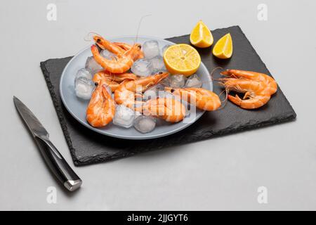 Garnelen, Zitrone und Eis auf einem grauen Teller und auf einer Steinoberfläche. Messer auf dem Tisch. Draufsicht. Grauer Hintergrund Stockfoto