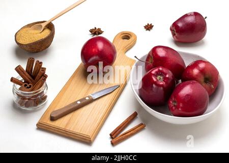 Reife rote Äpfel in einer Schüssel. Apfel und Messer auf Schneidebrett. Zimtstangen im Glasgefäß. Draufsicht. Weißer Hintergrund Stockfoto