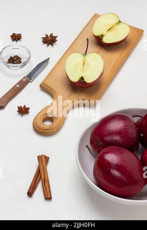 Apfelhälften auf Schneidebrett. Äpfel in einer Schüssel. Zimtschnecken, Sternanise und Messer auf dem Tisch. Draufsicht. Weißer Hintergrund Stockfoto