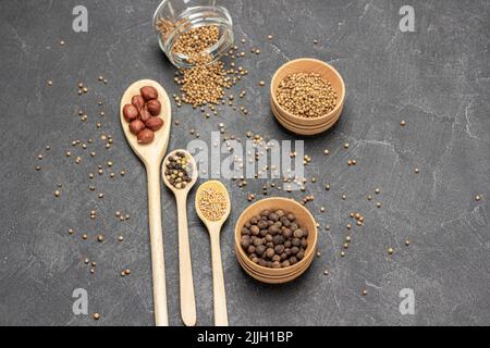 Gewürze in Holzlöffeln. Koriandersamen im Glasgefäß. Piment in Holzschale. Speicherplatz kopieren. Flach liegend. Schwarzer Hintergrund. Stockfoto