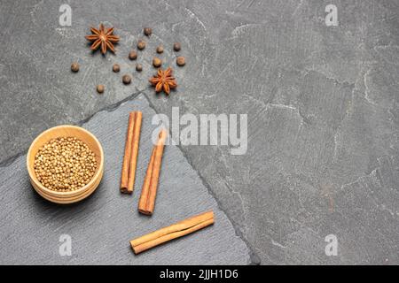 Koriandersamen in Holzschüssel und Zimtstangen auf Steinbrett. Sternanise und Piment auf dem Tisch. Speicherplatz kopieren. Draufsicht. Schwarzer Hintergrund Stockfoto