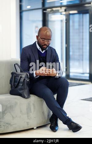 Ein afrikanischer Geschäftsmann tippt eine Nachricht auf sein Mobiltelefon, während er auf der Couch in der Lobby sitzt und auf das Treffen wartet Stockfoto