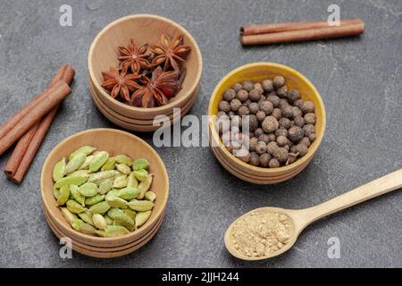 Piment, Kardamom und Sternanise in Schalen. Zimtstangen und trockene Kräuter in Holzlöffeln. Flach liegend. Schwarzer Hintergrund Stockfoto