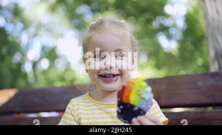 Kleines Mädchen hält einen bunten Lebkuchen und lächelt. Nahaufnahme Porträt von niedlichen Mädchen auf der Parkbank mit Cookies in sieht in die Ca sitzen Stockfoto