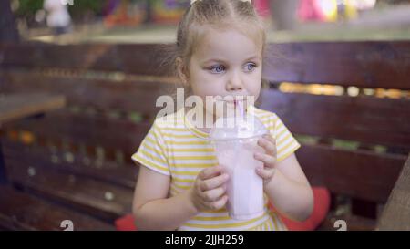 Das kleine Mädchen trinkt einen Milchshake durch einen Strohhalm. Nahaufnahme Porträt von niedlichen Mädchen auf Parkbank sitzen und trinken Milchshake Stockfoto