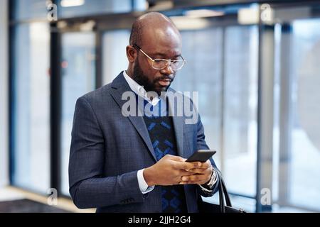 Seriöser afrikanischer Geschäftsmann im Anzug, der an seinem Mobiltelefon arbeitet und online mit seinem Kollegen spricht und eine Nachricht tippt Stockfoto