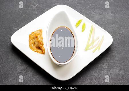 Gekochter Gyoza-Knödel und gehackter Lauch auf weißem Teller. Sojasauce in Porzellanschüssel. Draufsicht. Schwarzer Hintergrund Stockfoto