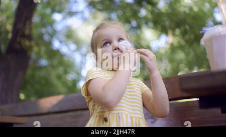 Odessa, Ukraine, Osteuropa. 26.. Juli 2022. Das kleine Mädchen wischt sich nach dem Essen mit einer Papierserviette ins Gesicht, auf dem Tisch liegt ein Milchshake. Nahaufnahme eines niedlichen Mädchen, das auf einer Parkbank sitzt und ihr Gesicht mit einer Papierserviette abwischt. (Bild: © Andrey Nekrasov/ZUMA Press Wire) Stockfoto