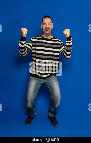 Ganzkörperbild eines sehr glücklichen und aufgeregt bärtigen hispanischen Mannes, der eine Siegergeste mit erhobenen Armen, einem Lächeln und einem Schreien nach Leistung vollbrachte. Stockfoto