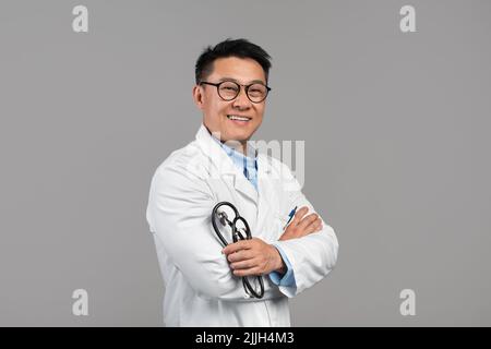 Lächelnd glücklich selbstbewusst Erwachsene koreanische Therapeut in weißem Mantel und Brille halten Thermometer, gekreuzte Arme auf der Brust Stockfoto