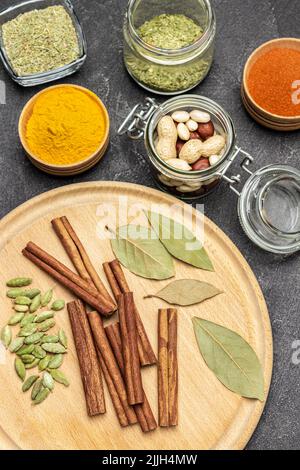 Zimtstangen, Kardamom und Lorbeerblatt auf Tafel. Kräuter und Nüsse in Gläsern trocknen. Kurkuma und Paprika in Schalen. Flach liegend. Schwarzer Hintergrund Stockfoto