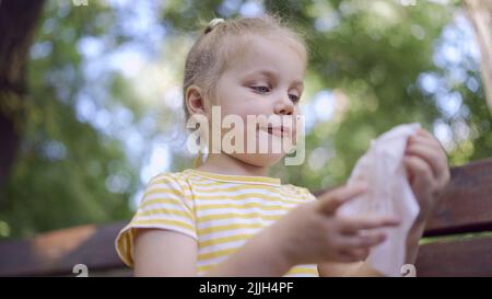 Odessa, Ukraine, Osteuropa. 26.. Juli 2022. Das kleine Mädchen wischt sich nach dem Essen mit einer Papierserviette ins Gesicht, auf dem Tisch liegt ein Milchshake. Nahaufnahme eines niedlichen Mädchen, das auf einer Parkbank sitzt und ihr Gesicht mit einer Papierserviette abwischt. (Bild: © Andrey Nekrasov/ZUMA Press Wire) Stockfoto