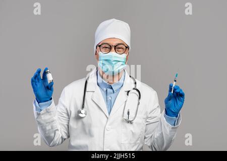 Fröhlicher reifer chinesischer Arzt in weißem Mantel, Brille und Handschuhen zeigt Impfstoff und Spritze Stockfoto