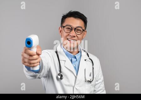 Happy adult asiatischen männlichen Arzt in weißem Mantel, Gläser misst Temperatur mit Infrarot-Thermometer Stockfoto