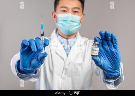 Seriöser erwachsener chinesischer Arzt mit weißem Mantel, Schutzmaske, Handschuhen und Spritze und Impfstoff Stockfoto