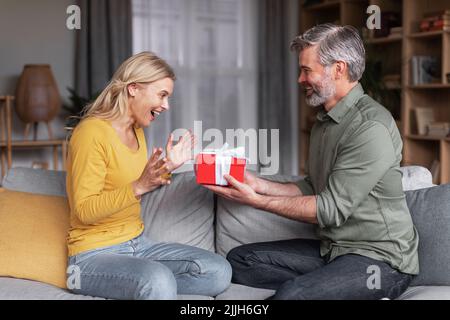 Liebevoller Mann mittleren Alters, der seine Frau mit einem Geschenk zu Hause überraschte Stockfoto