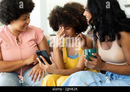 Fröhliche Biracial-Freundinnen necken junge Frau mit dem Smartphone, während sie zu Hause auf dem Sofa sitzen Stockfoto