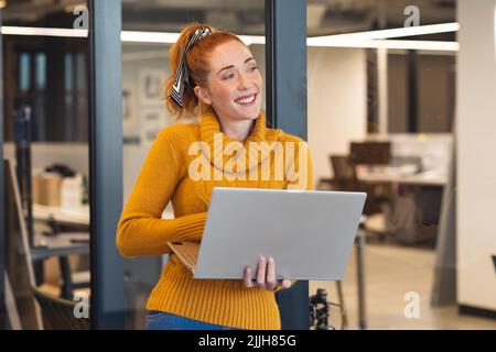 Lächelnde kaukasische Geschäftsfrau mit mittlerem Erwachsenenalter, die im Büro auf einem Laptop steht Stockfoto