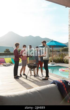 Multirassische männliche Freunde mit Bier und genießen Grillparty im Hof gegen Himmel im Sommer Stockfoto