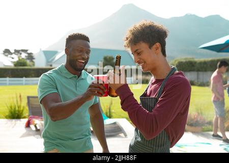 Fröhliche multirassische junge männliche Freunde, die Bier toasten, während sie sich auf der Sommerparty im Hof amüsieren Stockfoto