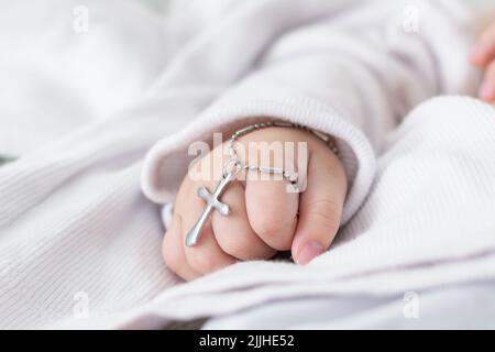 Nahaufnahme Detail der Hand eines Babys bei seiner Taufe, hält ein christliches Kreuz. katholische Taufe in lateinischer Kirche. kaukasisches Baby mit einem silbernen Kreuz clutc Stockfoto