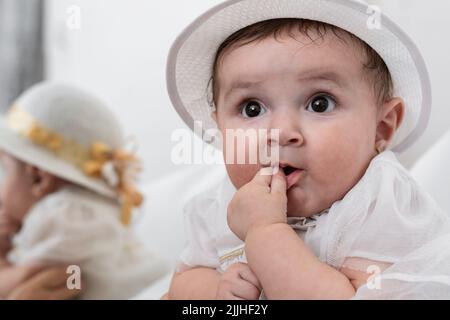 Nahaufnahme eines wunderschönen weißhäutigen Latina-Mädchens mit weißem Kleid und Hut, das mit Neugier und einem kleinen Lächeln ihre Mutter mit einem han anschaut Stockfoto