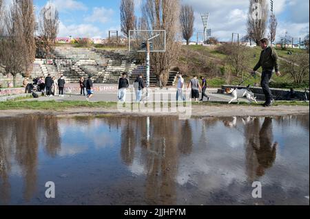 23.02.2022, Berlin, Deutschland, Europa - der Mensch geht mit seinem Hund und spiegelt sich in einer Pfütze im Mauerpark in Prenzlauer Berg, während junge Männer Basketball spielen. Stockfoto