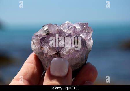 Schöne lila Amethyst Kristalle in einer weiblichen Hand. Druse Amethyst in Form eines Herzens auf dem Hintergrund des Meeres. Stockfoto