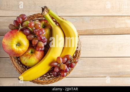 Reife Früchte im Weidenkorb auf Holzhintergrund. Bananen, Äpfel und Trauben. Erntekonzept. Speicherplatz kopieren. Stockfoto