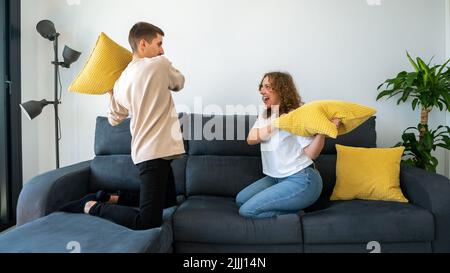 Ein paar von zwei glücklichen Freunden, die Spaß haben, zusammen auf dem Sofa zu Hause zu spielen und mit Kissen zu kämpfen genießen. Die Kissen waren morgens drinnen Stockfoto