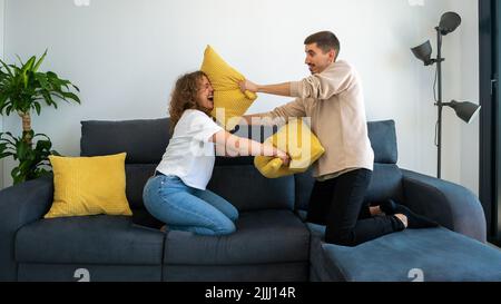 Ein paar von zwei glücklichen Freunden, die Spaß haben, zusammen auf dem Sofa zu Hause zu spielen und mit Kissen zu kämpfen genießen. Die Kissen waren morgens drinnen Stockfoto