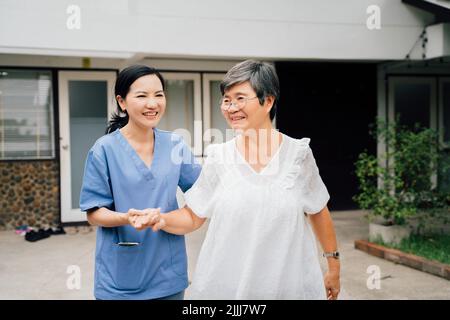 Optimistische Krankenschwester in blauer Uniform, die eine ältere asiatische Frau unterstützt, während sie gemeinsam durch das Tor vor dem Haus geht Stockfoto