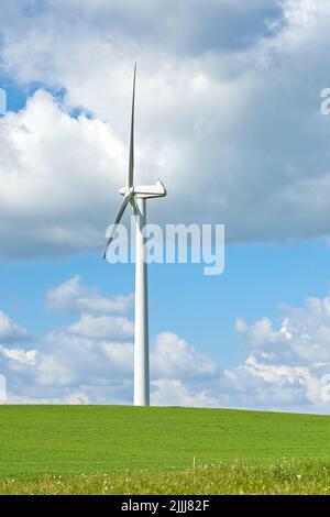 Windmühle. Eine Windmühle auf einem grünen Hügel gegen wolkigen blauen Himmel mit Kopierraum. Windturbinen erzeugen Strom durch sich drehende Propellerblätter Stockfoto